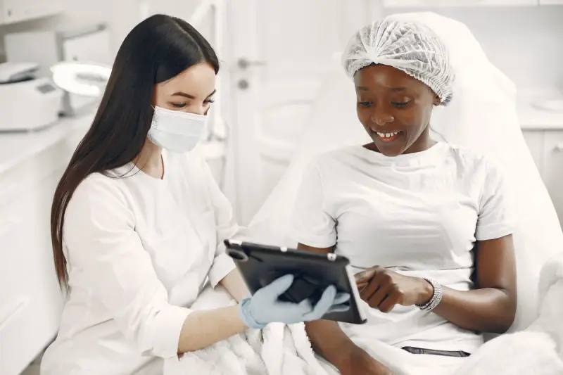 Masked clinician in gloves shows a smiling patient results on a tablet in a bright treatment room