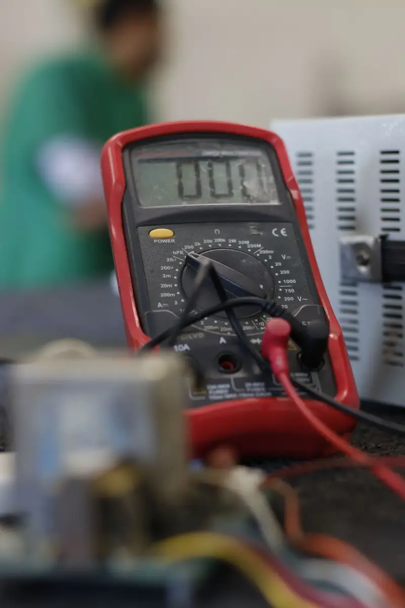 Red digital multimeter with test leads on a repair bench beside an electronics enclosure
