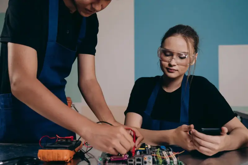 Two technicians in work aprons probe a circuit board with multimeter leads at a workbench