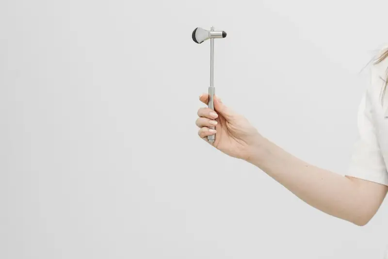 Hand of a clinician holding a steel reflex hammer against a plain light background