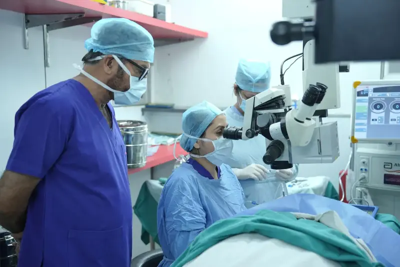 Woman eye surgeon looks through an operating microscope while a colleague watches beside a monitor