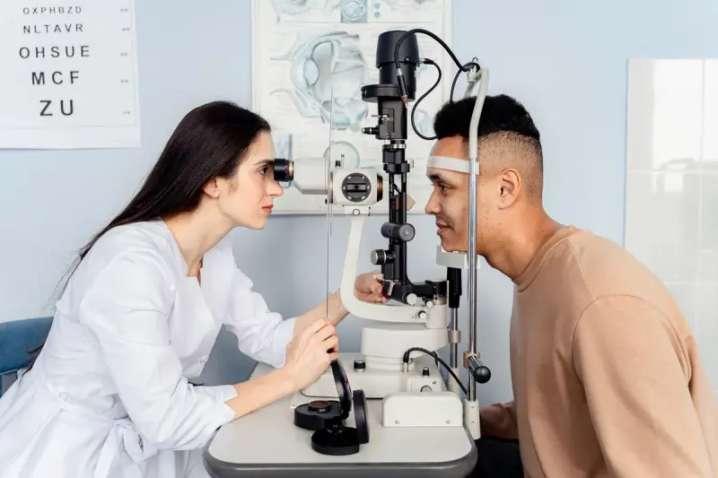 Woman doctor in a white coat examines a seated man's eyes with a slit lamp, eye chart behind