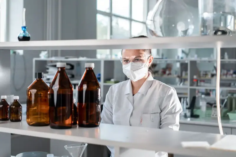 Female scientist in goggles and mask behind a shelf of amber chemical bottles in a lab