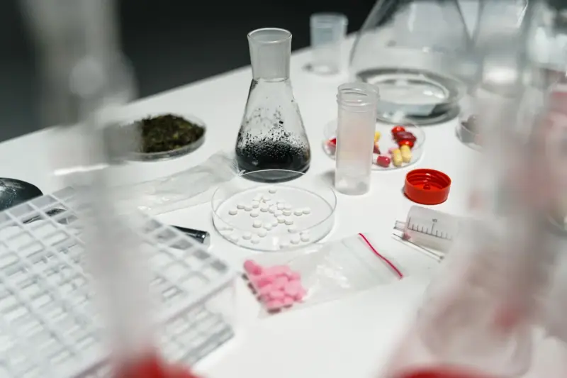 Tablets in a glass dish among flasks, vials and capsules on a white lab table