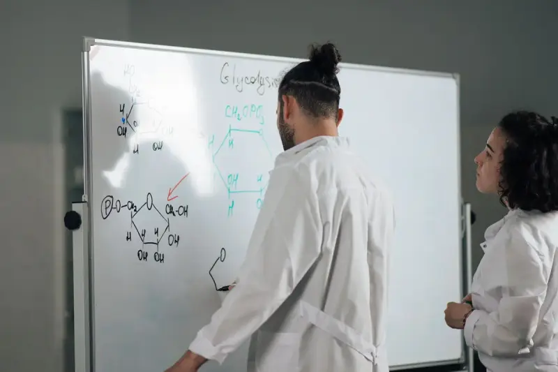 Two scientists in lab coats working through chemical structures on a whiteboard