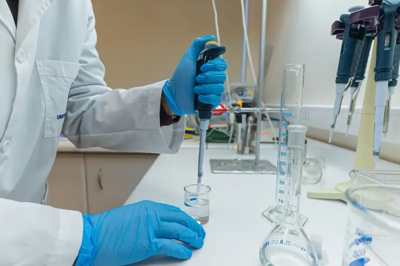 Scientist in a lab coat and blue gloves pipetting liquid into a small beaker