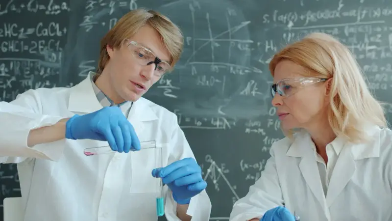 Two scientists in goggles and gloves comparing a test tube in front of a chalkboard of equations