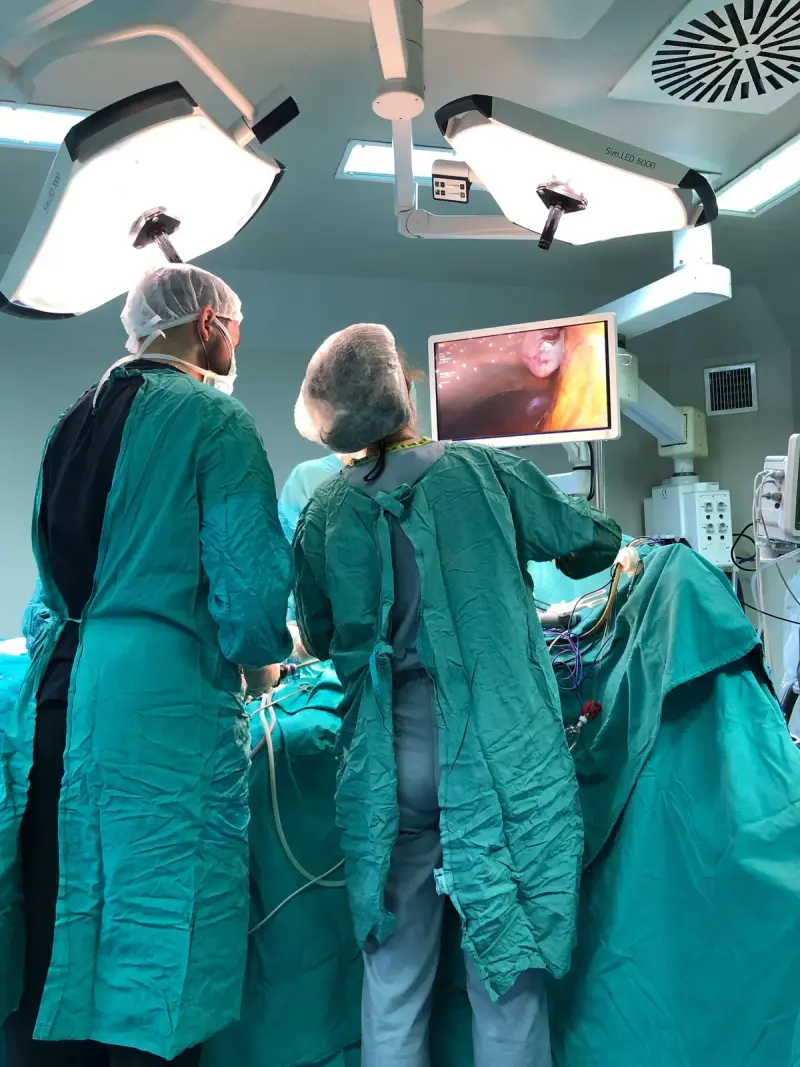 A woman and a man in green gowns operating while watching a camera monitor under surgical lights