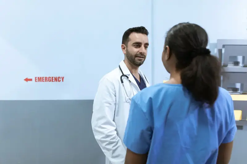 Physician in a white coat talking with a nurse in blue scrubs beside an emergency department sign