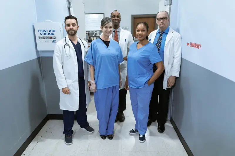 Five doctors and nurses in white coats and scrubs smiling together in a hospital hallway