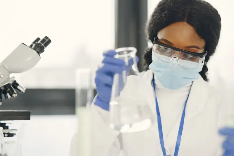 Chemist in goggles and mask holding a flask beside a microscope