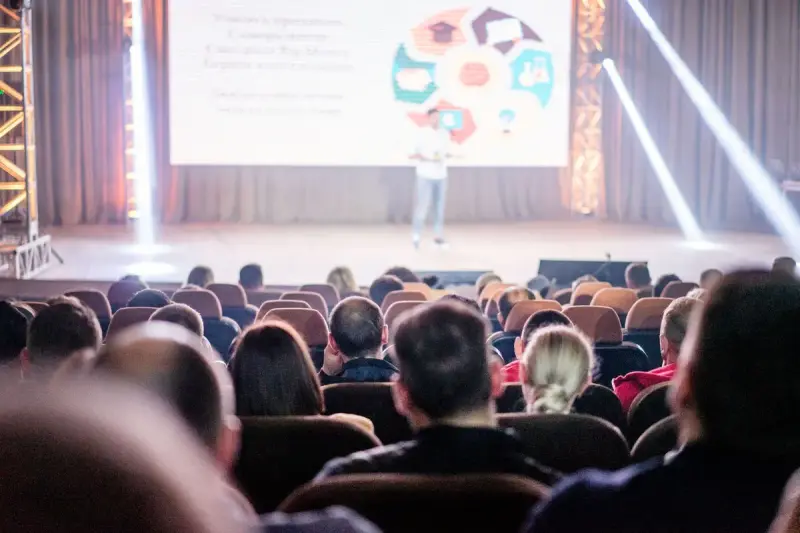 Audience seated in a darkened auditorium watching a speaker present on stage