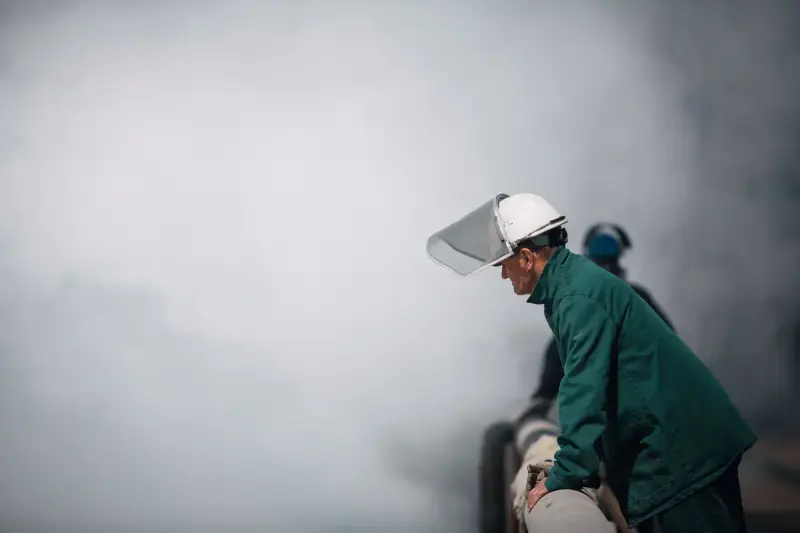 Worker in a face shield and hard hat beside a cloud of steam
