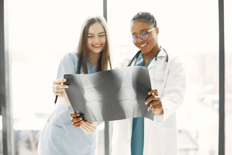 Two smiling clinicians in scrubs and a white coat reviewing an X-ray film by a window