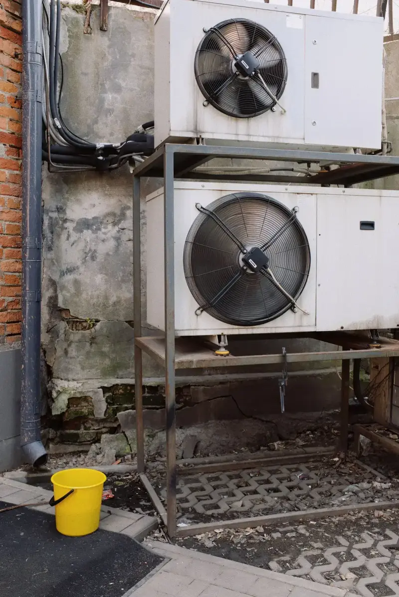 Two outdoor condenser units stacked on a steel frame beside a brick wall