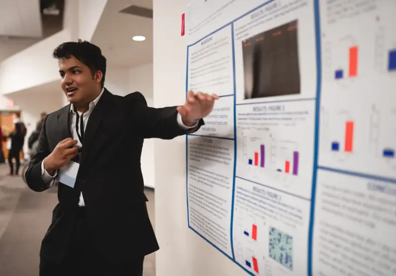 Young researcher in a dark suit explaining results on a printed scientific poster
