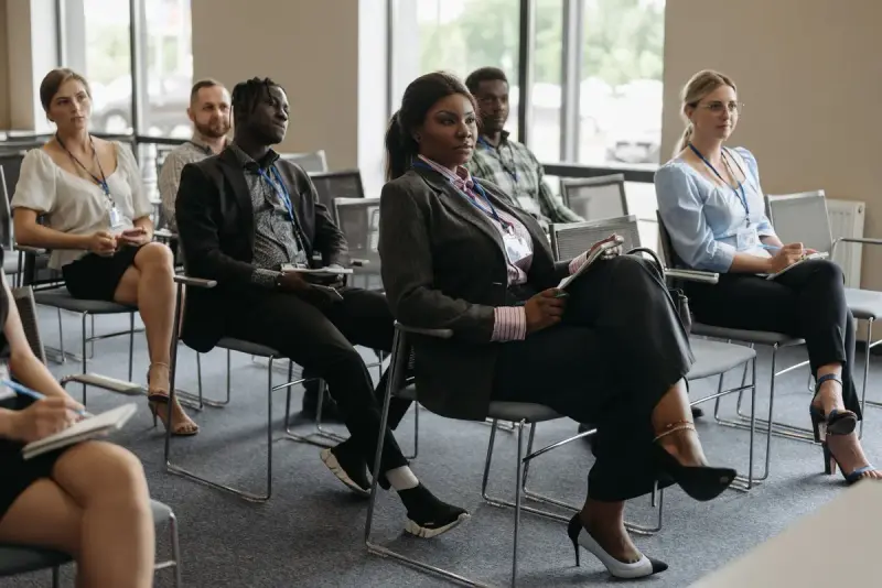 Professionals with lanyards and notebooks listening to a speaker in a bright meeting room