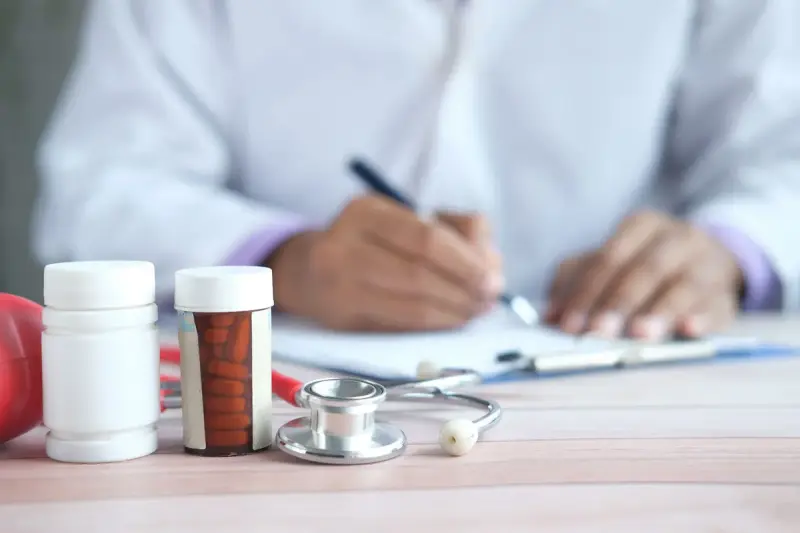 Hand writing on a form beside pill bottles and a stethoscope on a desk