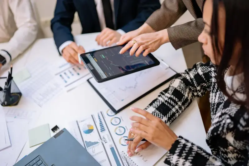 Businesswoman reviewing charts on a tablet with colleagues around a table of reports
