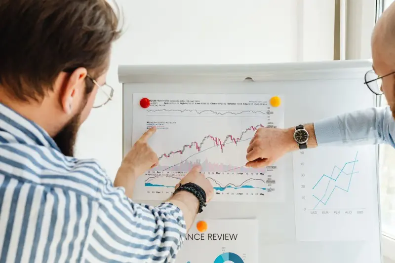 Two men discussing a line chart pinned to a whiteboard in a bright office