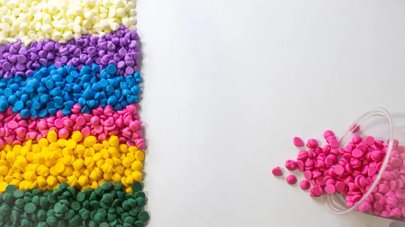Bands of white, purple, blue, pink, yellow and green plastic pellets beside a cup of spilled pink resin