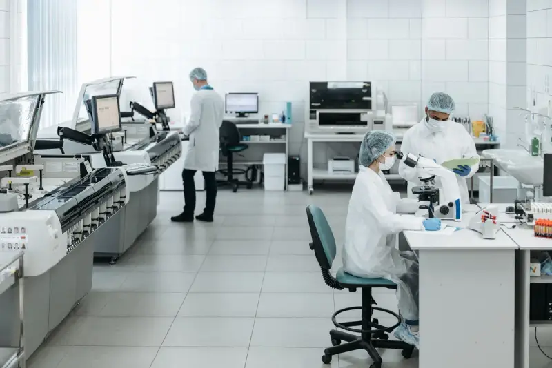 Clinical laboratory with automated analyzers along one wall and three scientists in caps and gowns at work