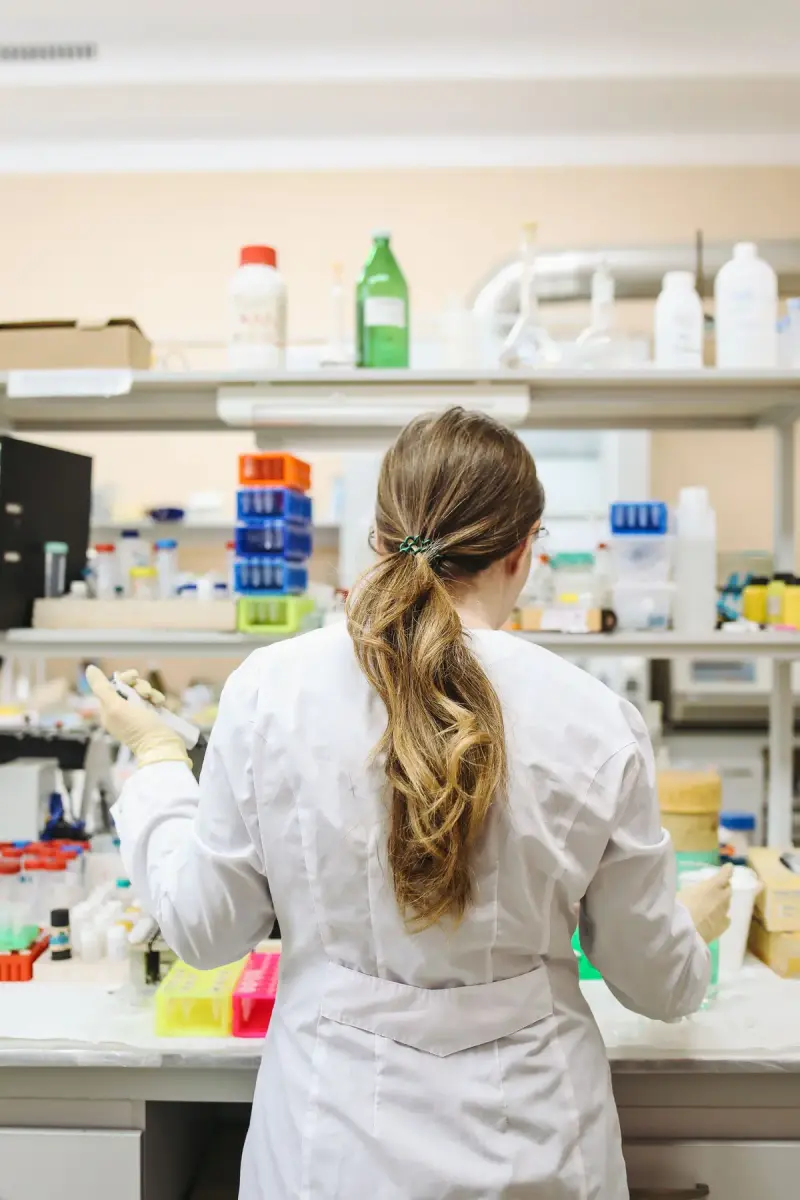 Woman in a lab coat working at a bench lined with tube racks and reagent bottles