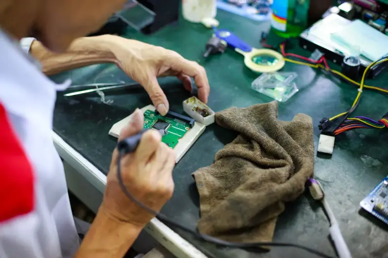 Technician's hands working on a small circuit board with a soldering iron at a cluttered bench