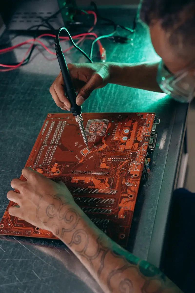 Engineer with tattooed arms soldering components onto a motherboard in a tech workshop