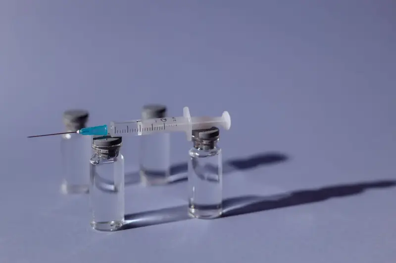 Syringe resting across three small glass vaccine vials on a lilac background