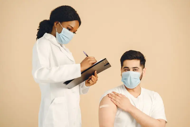 Masked clinician with a clipboard beside a patient with a bandage after a vaccination