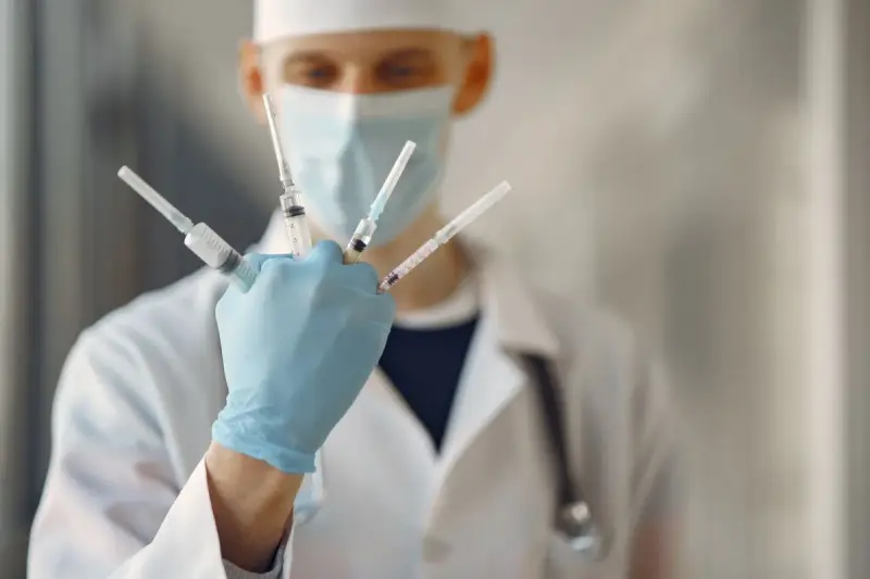 Masked clinician in a white coat and gloves holding four prepared syringes