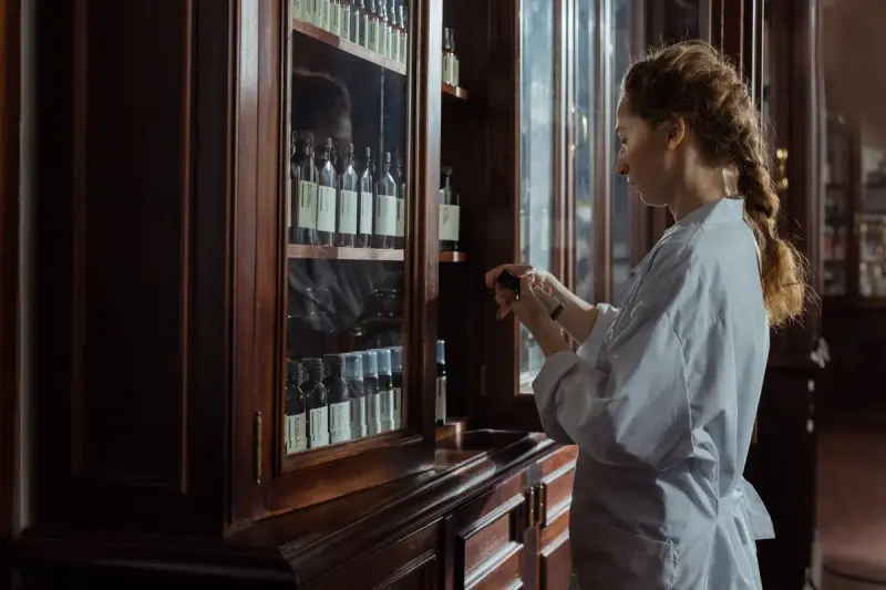 Pharmacist in a white coat checking a bottle from a wooden pharmacy cabinet