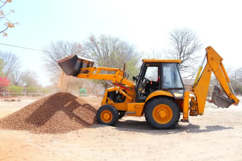 Yellow backhoe loader dumping a bucket of gravel onto a pile on a sunny site