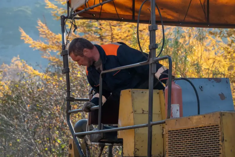 Operator in blue workwear at the controls of a compact machine with autumn trees behind