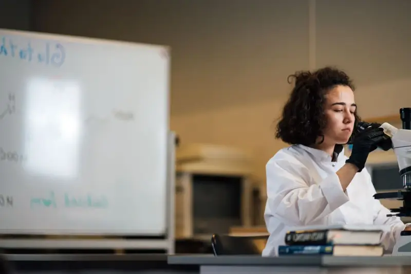 Female scientist in a lab coat and gloves looking into a microscope