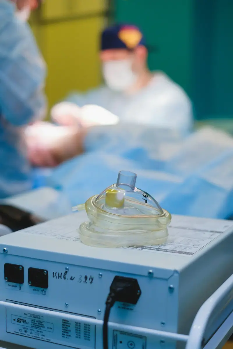 Clear anesthesia face mask resting on a device in an operating room, clinicians blurred behind