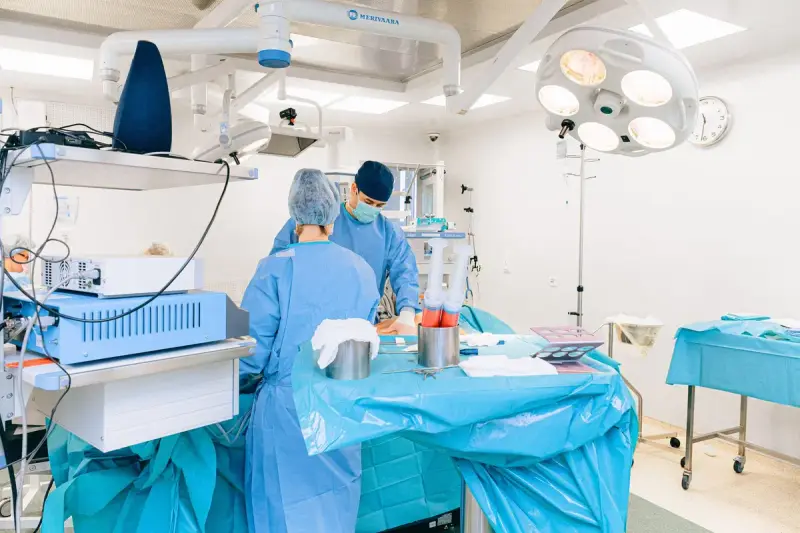Two clinicians in blue gowns working at a draped table beside equipment carts and surgical lights