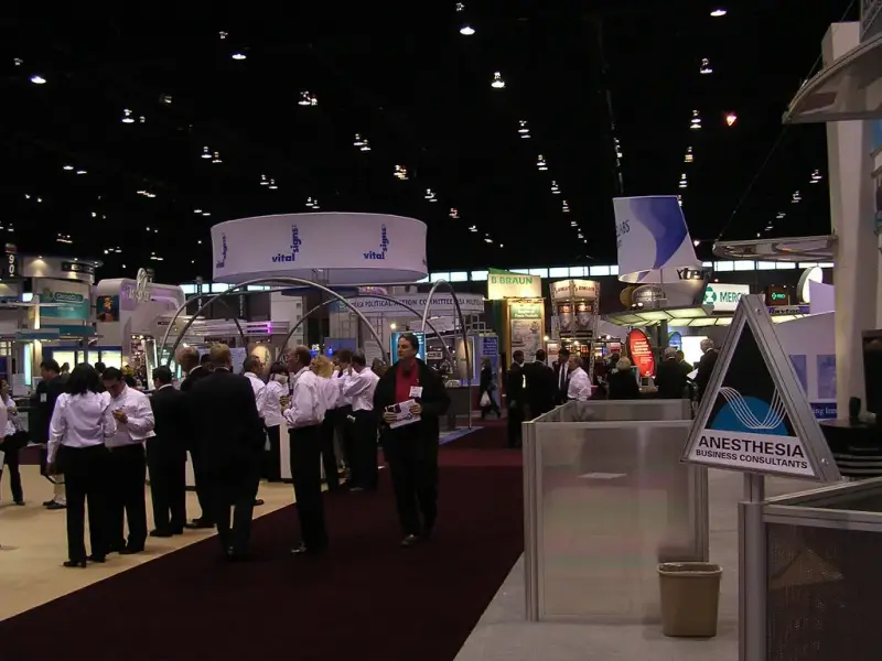 Attendees walking a carpeted aisle between exhibits under a dark exhibit hall ceiling
