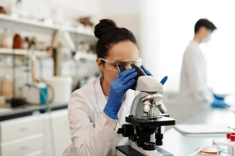 Researcher in safety glasses at a microscope in a bright lab while a colleague works at the bench behind her