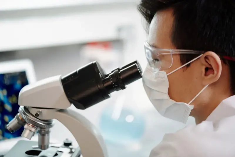 Scientist in a face mask and safety glasses looking through a microscope in a white lab