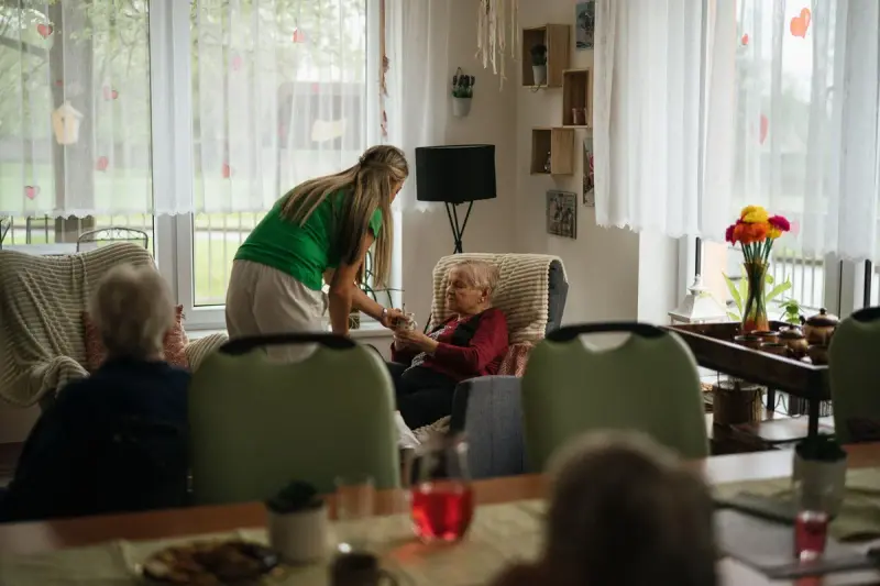 Caregiver in green leaning in to help older residents at a table in a bright care home lounge
