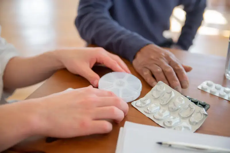 Two people sorting tablets from blister packs into a round pill box on a table