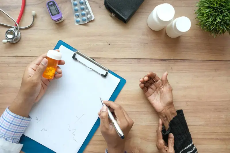 Clinician holding an orange pill bottle and clipboard across a desk from an older patient