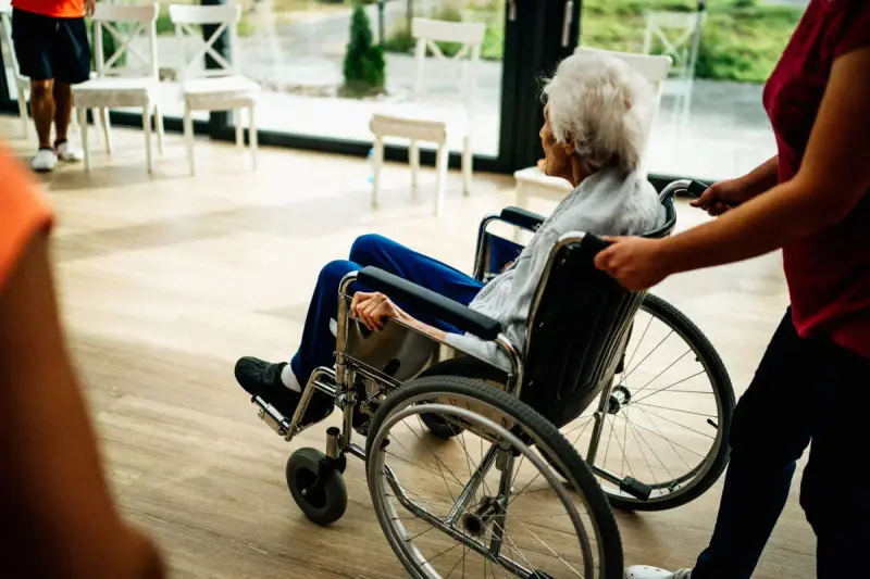 Care worker pushing an older woman in a wheelchair toward a sunny window
