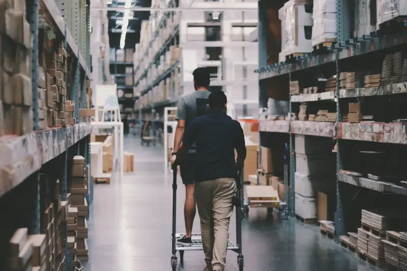 Two men pushing a flat trolley down a tall warehouse aisle lined with boxed merchandise