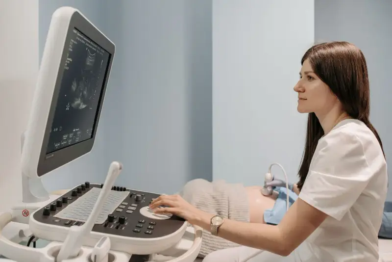 Sonographer in white scrubs runs an ultrasound exam at a cart-based console in a pale blue exam room