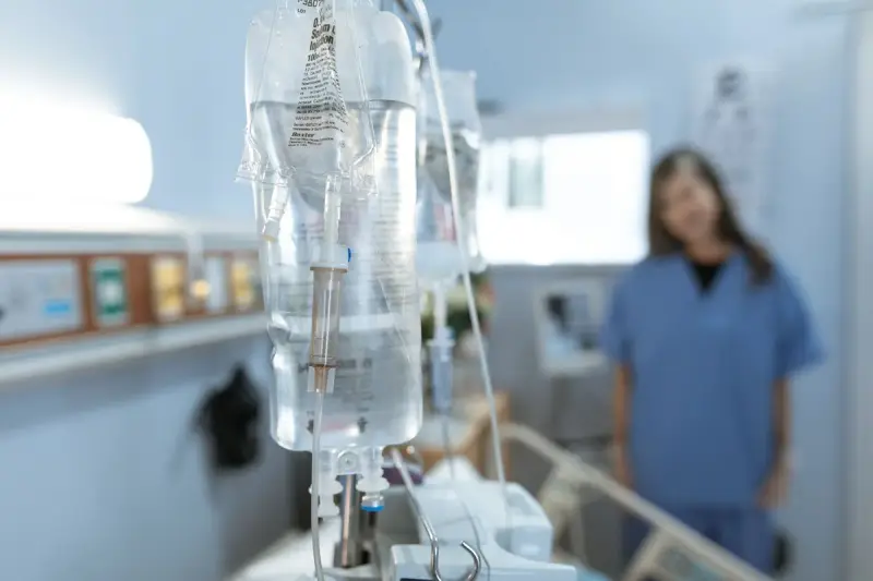 IV bags hanging over a hospital bed with a nurse in blue scrubs in the background