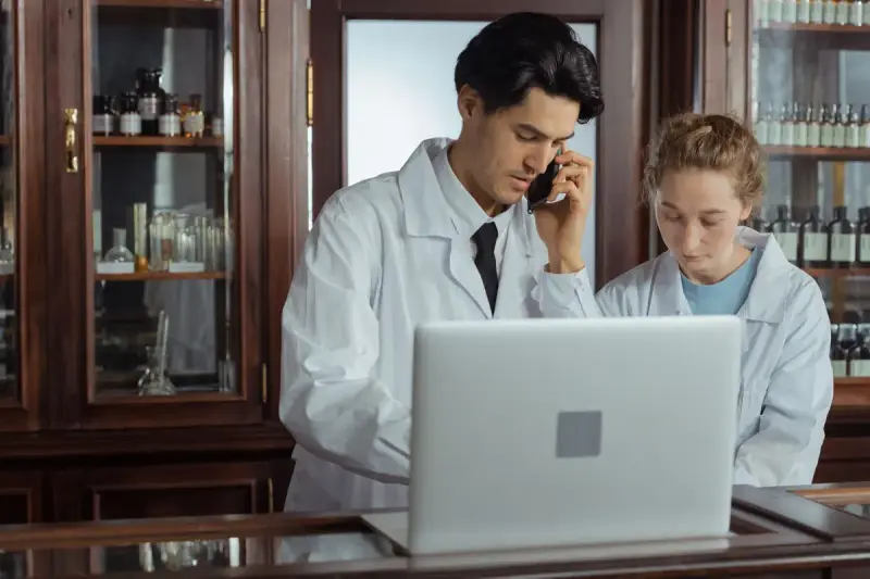 Two pharmacists in white coats at a counter with a laptop, one on the phone