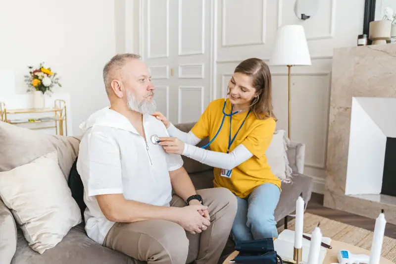 Nurse in a yellow shirt listening to an older man's chest with a stethoscope at home